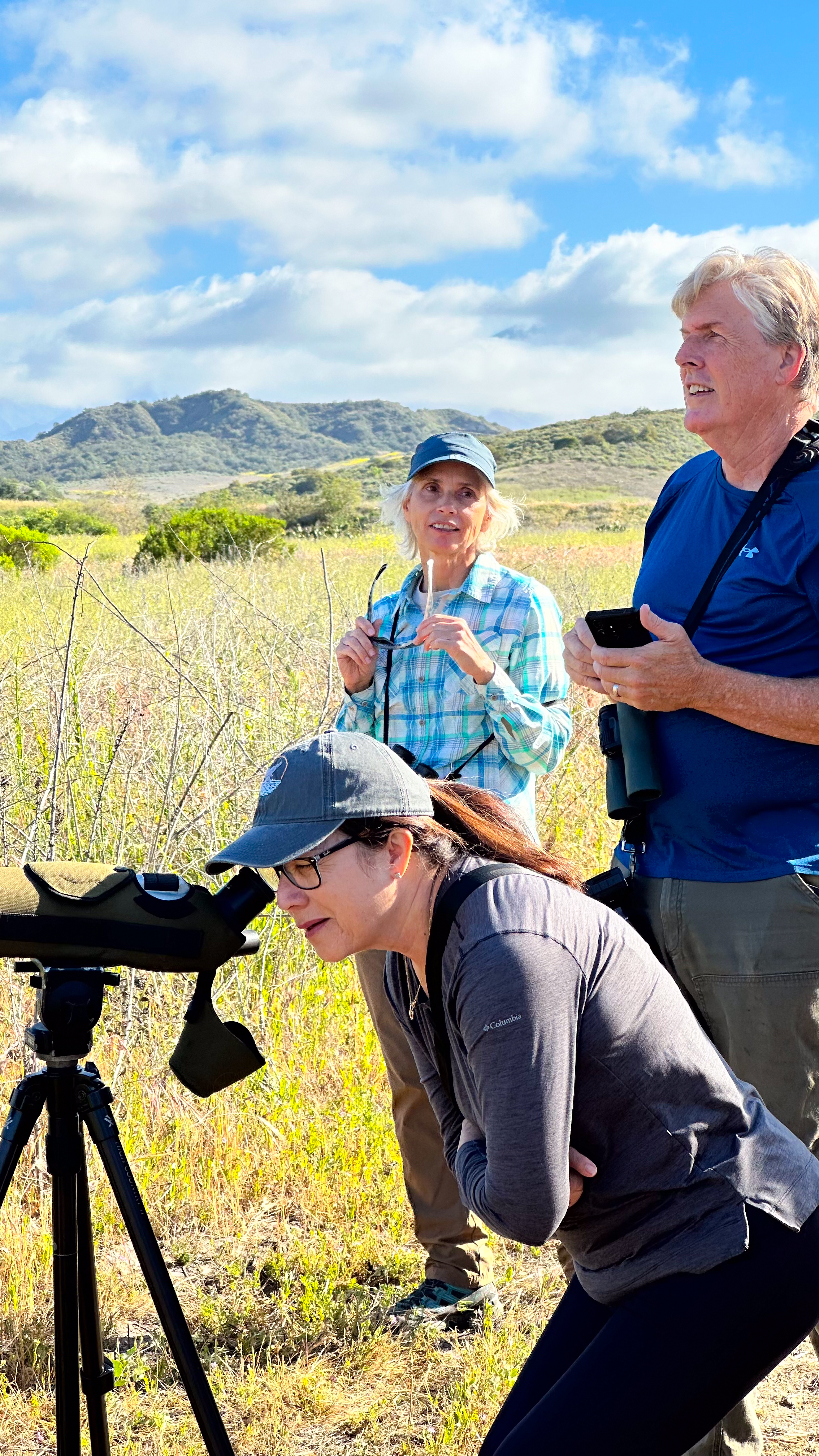 a person looking through binoculars at wildlife in a grassy field with hills and two other people standing in the background